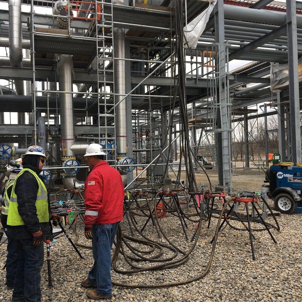 Two crew members at a test manifold with hose bundles under plant scaffolding